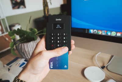 A hand holding a card reader and a blue Visa card in front of a desk workspace that includes a monitor displaying a blue screen. There is a decorative plant in a pot, a partially visible package, and a wireless charger on the desk.