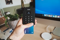 A hand holding a card reader and a blue Visa card in front of a desk workspace that includes a monitor displaying a blue screen. There is a decorative plant in a pot, a partially visible package, and a wireless charger on the desk.