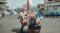 A street vendor is seen pushing a cart filled with various food items and supplies. The cart is colorful and packed with bottles, boxes, and bags. The vendor is wearing a casual t-shirt and appears to be in motion on a city street, which is bustling with other people and vehicles. Buildings and urban infrastructure are visible in the background.