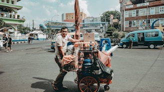 A street vendor is seen pushing a cart filled with various food items and supplies. The cart is colorful and packed with bottles, boxes, and bags. The vendor is wearing a casual t-shirt and appears to be in motion on a city street, which is bustling with other people and vehicles. Buildings and urban infrastructure are visible in the background.