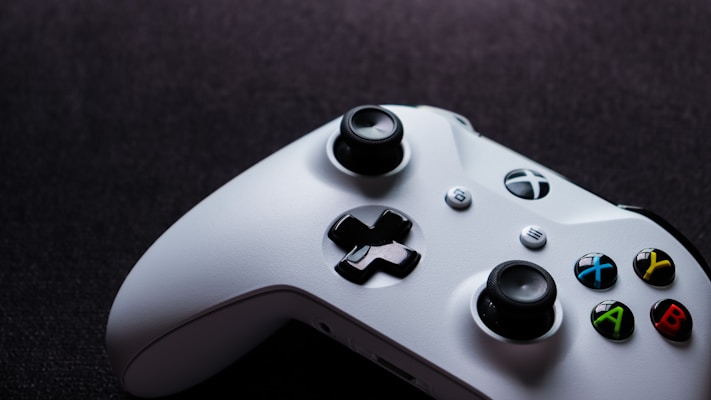 A white gaming controller with black buttons and colorful accents, including green, red, yellow, and blue buttons, is placed on a dark, textured surface. The design features dual analog sticks, a directional pad, and various control buttons, some labeled with letters.