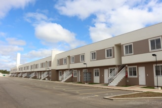 A row of modern, two-story townhouses with beige and white exteriors, featuring multiple windows and white staircases leading to the upper level. A spacious, empty parking lot is in the foreground, and the sky is blue with scattered clouds.