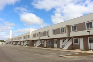 A row of modern, two-story townhouses with beige and white exteriors, featuring multiple windows and white staircases leading to the upper level. A spacious, empty parking lot is in the foreground, and the sky is blue with scattered clouds.