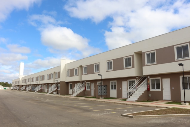 A row of modern, two-story townhouses with beige and white exteriors, featuring multiple windows and white staircases leading to the upper level. A spacious, empty parking lot is in the foreground, and the sky is blue with scattered clouds.