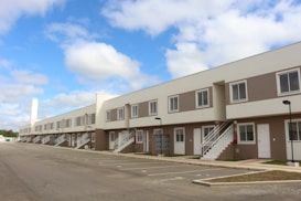 A row of modern, two-story townhouses with beige and white exteriors, featuring multiple windows and white staircases leading to the upper level. A spacious, empty parking lot is in the foreground, and the sky is blue with scattered clouds.