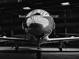 A sleek, reflective aircraft nose is housed inside a dimly lit hangar. The reflection of the hangar ceiling is clearly visible on the polished surface of the plane. The lighting creates a dramatic contrast, highlighting the aircraft against the shadowy background.