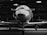 A sleek, reflective aircraft nose is housed inside a dimly lit hangar. The reflection of the hangar ceiling is clearly visible on the polished surface of the plane. The lighting creates a dramatic contrast, highlighting the aircraft against the shadowy background.