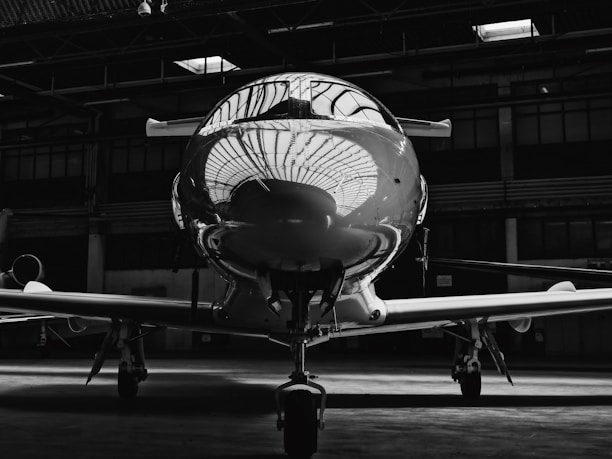 A sleek, reflective aircraft nose is housed inside a dimly lit hangar. The reflection of the hangar ceiling is clearly visible on the polished surface of the plane. The lighting creates a dramatic contrast, highlighting the aircraft against the shadowy background.