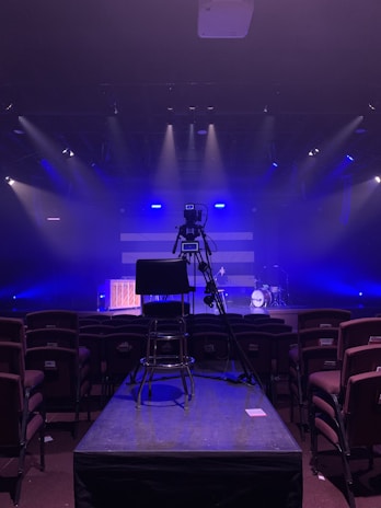 An empty stage setup featuring a camera on a tripod facing a piano and a drum kit under dim, blue lighting. Rows of empty chairs are arranged in the foreground, suggesting a concert or event venue.