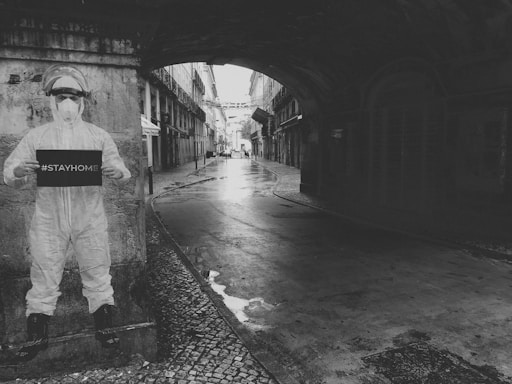 A person in full protective gear, including a mask and face shield, stands near a wall holding a sign that reads '#STAYHOME'. The setting includes a narrow, empty street framed by tall buildings, leading into an archway. The overall atmosphere is quiet and somewhat somber, with wet cobblestones suggesting recent rain.