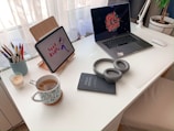 A tidy white desk with a laptop and a tablet. The laptop screen displays 'get it done' and the tablet shows 'good karma' with an illustration. On the desk, there are headphones, a notebook labeled 'FIELD NOTES', a cup of tea with a floral pattern mug, an assortment of colorful pens in a holder, and a lit candle. A potted plant is visible in the corner next to white curtains.