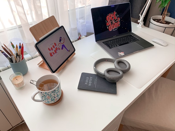 A tidy white desk with a laptop and a tablet. The laptop screen displays 'get it done' and the tablet shows 'good karma' with an illustration. On the desk, there are headphones, a notebook labeled 'FIELD NOTES', a cup of tea with a floral pattern mug, an assortment of colorful pens in a holder, and a lit candle. A potted plant is visible in the corner next to white curtains.