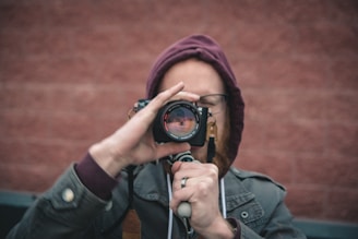 A person holds a camera up to their eye with both hands, focusing as they take a photograph. The individual is wearing a maroon hood and a gray jacket, standing in front of a textured, red brick wall.