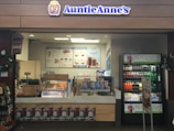 A retail kiosk featuring Auntie Anne's branding is filled with displays of pretzel snacks and beverages. Various drinks are visible in a refrigerated section, and promotional signs highlight new product offerings such as pretzel bundles and chocolate frost drinks. Decor includes wreaths, and the counter has a cash register for processing purchases.