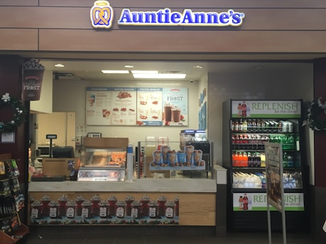 A retail kiosk featuring Auntie Anne's branding is filled with displays of pretzel snacks and beverages. Various drinks are visible in a refrigerated section, and promotional signs highlight new product offerings such as pretzel bundles and chocolate frost drinks. Decor includes wreaths, and the counter has a cash register for processing purchases.