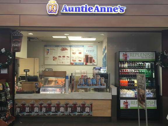 A retail kiosk featuring Auntie Anne's branding is filled with displays of pretzel snacks and beverages. Various drinks are visible in a refrigerated section, and promotional signs highlight new product offerings such as pretzel bundles and chocolate frost drinks. Decor includes wreaths, and the counter has a cash register for processing purchases.
