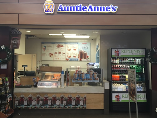 A retail kiosk featuring Auntie Anne's branding is filled with displays of pretzel snacks and beverages. Various drinks are visible in a refrigerated section, and promotional signs highlight new product offerings such as pretzel bundles and chocolate frost drinks. Decor includes wreaths, and the counter has a cash register for processing purchases.