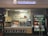 A retail kiosk featuring Auntie Anne's branding is filled with displays of pretzel snacks and beverages. Various drinks are visible in a refrigerated section, and promotional signs highlight new product offerings such as pretzel bundles and chocolate frost drinks. Decor includes wreaths, and the counter has a cash register for processing purchases.