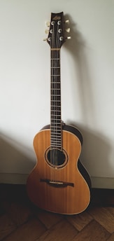 A wooden acoustic guitar with six strings is positioned against a plain wall. The guitar features a light brown body with darker edges, a round sound hole with decorative rosette, and a dark fretboard extending up to a headstock with six tuning pegs.