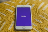 A smartphone with a white frame displays the Zelle app on its screen, which features a purple background. The phone is placed on a yellow crocheted fabric, adding texture to the background.