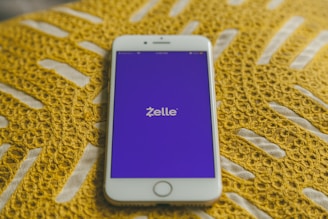 A smartphone with a white frame displays the Zelle app on its screen, which features a purple background. The phone is placed on a yellow crocheted fabric, adding texture to the background.