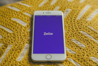 A smartphone with a white frame displays the Zelle app on its screen, which features a purple background. The phone is placed on a yellow crocheted fabric, adding texture to the background.