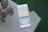 A smartphone is placed on a checkered surface outdoors, displaying a payment or request app where someone is trying to send or request $100 for utilities. There is a potted plant with succulent leaves in the corner.