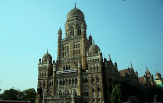A grand, historic building with elaborate architecture featuring domes, arched windows, and ornate details. It is constructed primarily from red brick, with contrasting white accents, and displays a blend of Gothic and Indo-Saracenic architectural styles. The structure is surrounded by lush greenery and set against a clear blue sky.