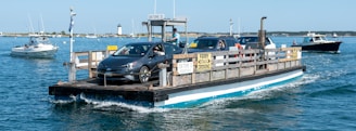 A small ferry is transporting cars across a body of water. The ferry appears to be in a coastal setting, with other boats visible in the background. Clear skies and calm blue water suggest a serene environment.