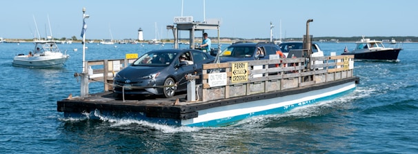 A small ferry is transporting cars across a body of water. The ferry appears to be in a coastal setting, with other boats visible in the background. Clear skies and calm blue water suggest a serene environment.