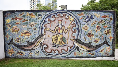 A vibrant mosaic artwork depicting a central figure resembling a sea god holding a trident. Surrounding the figure are various marine animals, including fish, dolphins, jellyfish, and starfish. The background is richly colored with blues and greens, adding to the aquatic theme. The mural is framed by a wave-like pattern, adding dynamic movement to the scene.