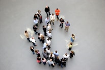 A group of people stand seemingly arranged in a circular formation on a light gray floor. Most individuals are casually dressed, with some appearing to engage in conversation or looking at something in their hands. The scene is viewed from above, creating a dynamic visual of human interaction.