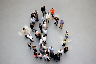 A group of people stand seemingly arranged in a circular formation on a light gray floor. Most individuals are casually dressed, with some appearing to engage in conversation or looking at something in their hands. The scene is viewed from above, creating a dynamic visual of human interaction.