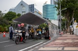 Several motorcyclists are riding through a tent-like structure on a city street. They are wearing helmets, and two uniformed officers are monitoring the scene. High-rise buildings and trees are visible in the background, and orange traffic cones line the street.