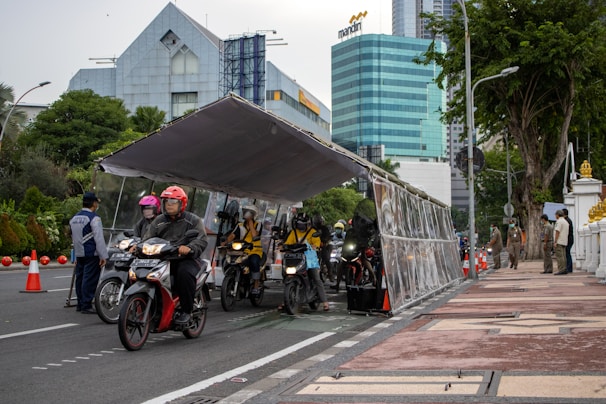 Several motorcyclists are riding through a tent-like structure on a city street. They are wearing helmets, and two uniformed officers are monitoring the scene. High-rise buildings and trees are visible in the background, and orange traffic cones line the street.