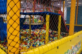 A children's indoor play area is filled with an array of colorful plastic balls enclosed in a mesh fence. The structure includes climbing features and slides, with various vibrant colors used throughout the setup.