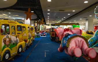 A colorful indoor amusement ride area with children’s mini rides, including school bus-themed rides and a pink elephant ride. The area is decorated with stars hanging from the ceiling and has a soft blue carpet.