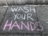 Brightly colored chalk writing on a concrete sidewalk with the words 'WASH YOUR HANDS' emphasized in white and pink.
