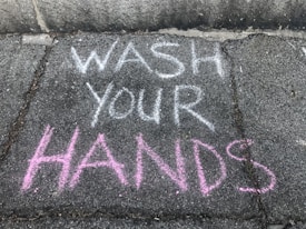 Brightly colored chalk writing on a concrete sidewalk with the words 'WASH YOUR HANDS' emphasized in white and pink.