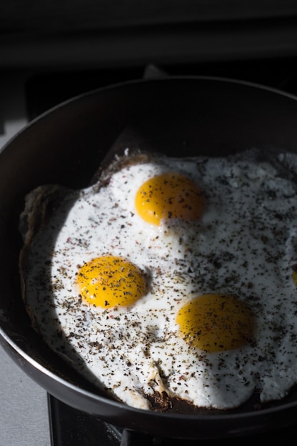 Three sunny-side-up eggs are cooking in a black frying pan, with some herbs and spices sprinkled on top. The eggs have bright yellow yolks and are surrounded by white egg whites.