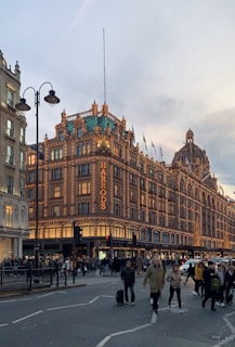 A large, iconic department store building is adorned with numerous decorative lights, creating a warm and inviting glow. The architecture is grand and classical, featuring a series of windows and ornate design elements. People are walking along the street in front, some carrying shopping bags, suggesting a busy urban environment. The sky is slightly overcast, hinting at evening or late afternoon.