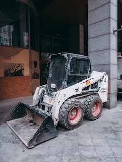 A compact skid-steer loader is parked on a gray tiled surface near a concrete column. The loader has white and black coloring with red wheels and a visible front bucket attachment. The surrounding area appears to be an urban environment, with buildings partially visible in the background.