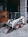 A compact skid-steer loader is parked on a gray tiled surface near a concrete column. The loader has white and black coloring with red wheels and a visible front bucket attachment. The surrounding area appears to be an urban environment, with buildings partially visible in the background.