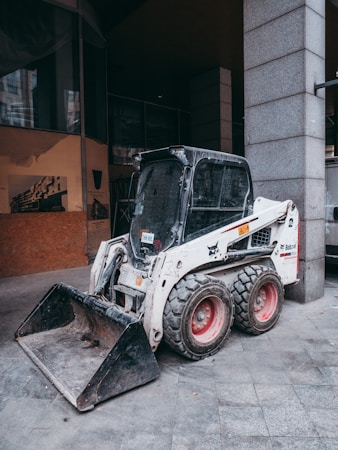 A compact skid-steer loader is parked on a gray tiled surface near a concrete column. The loader has white and black coloring with red wheels and a visible front bucket attachment. The surrounding area appears to be an urban environment, with buildings partially visible in the background.