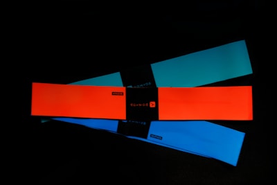 Three elastic resistance bands are laid out on a dark surface. The bands are rectangular and come in different colors: bright orange, teal, and blue. Each band has a black label in the center with branding or text.