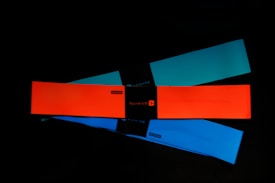 Three elastic resistance bands are laid out on a dark surface. The bands are rectangular and come in different colors: bright orange, teal, and blue. Each band has a black label in the center with branding or text.