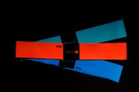 Three elastic resistance bands are laid out on a dark surface. The bands are rectangular and come in different colors: bright orange, teal, and blue. Each band has a black label in the center with branding or text.
