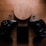 A wooden wireless charger is placed on a wooden surface, surrounded by two black gaming controllers and a small black electronic device. The setup includes two black thumb grips and emits a warm, moody ambiance.