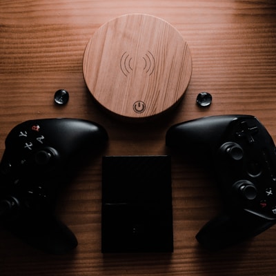 A wooden wireless charger is placed on a wooden surface, surrounded by two black gaming controllers and a small black electronic device. The setup includes two black thumb grips and emits a warm, moody ambiance.