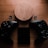 A wooden wireless charger is placed on a wooden surface, surrounded by two black gaming controllers and a small black electronic device. The setup includes two black thumb grips and emits a warm, moody ambiance.